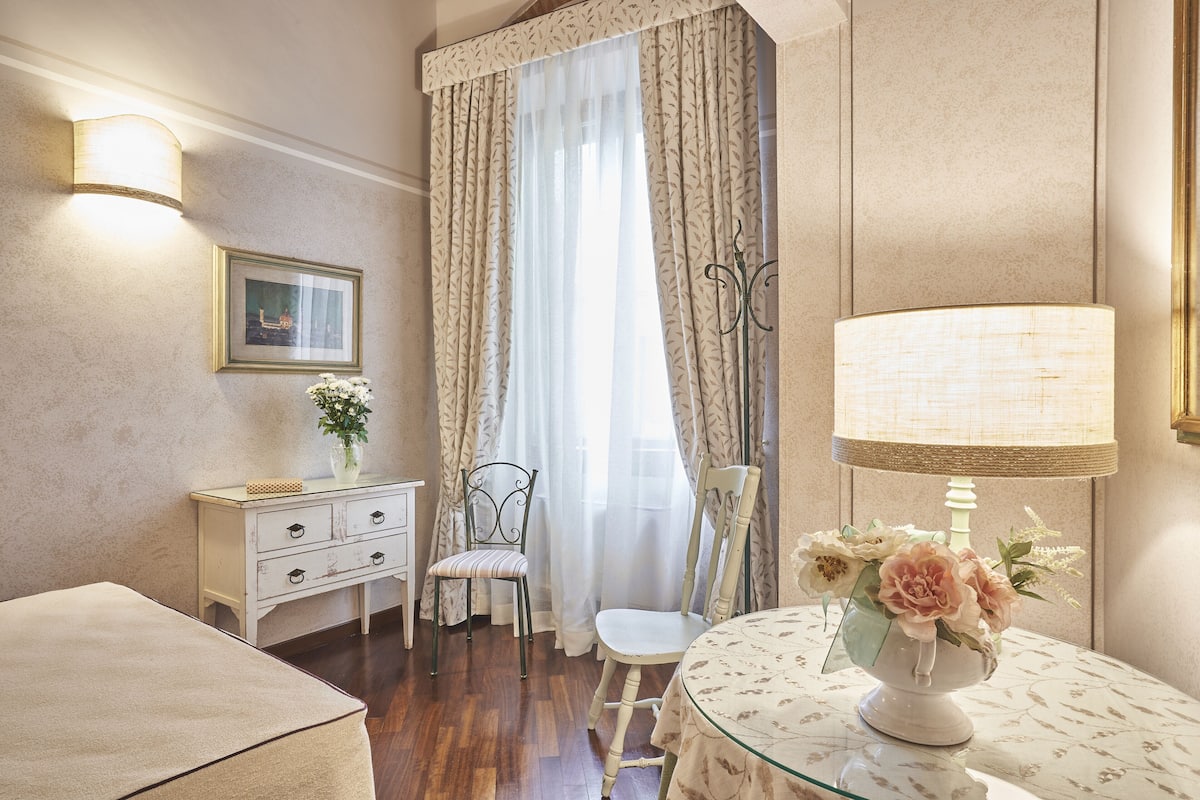 A room with a wooden floor, a white sofa, a small table with a vase of flowers, a lamp, a chair, a dresser, and a window with sheer curtains.