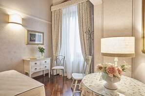 Ein Zimmer mit Holzboden, einem weißen Sofa, einem kleinen Tisch mit Blumenvase, einer Lampe, einem Sessel, einer Kommode und einem Fenster mit durchsichtigen Vorhängen.