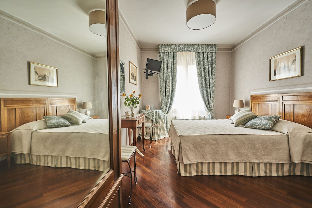 A hotel room with two beds, a wooden headboard, a desk with a chair, a small table with a vase of flowers, and a large mirror.