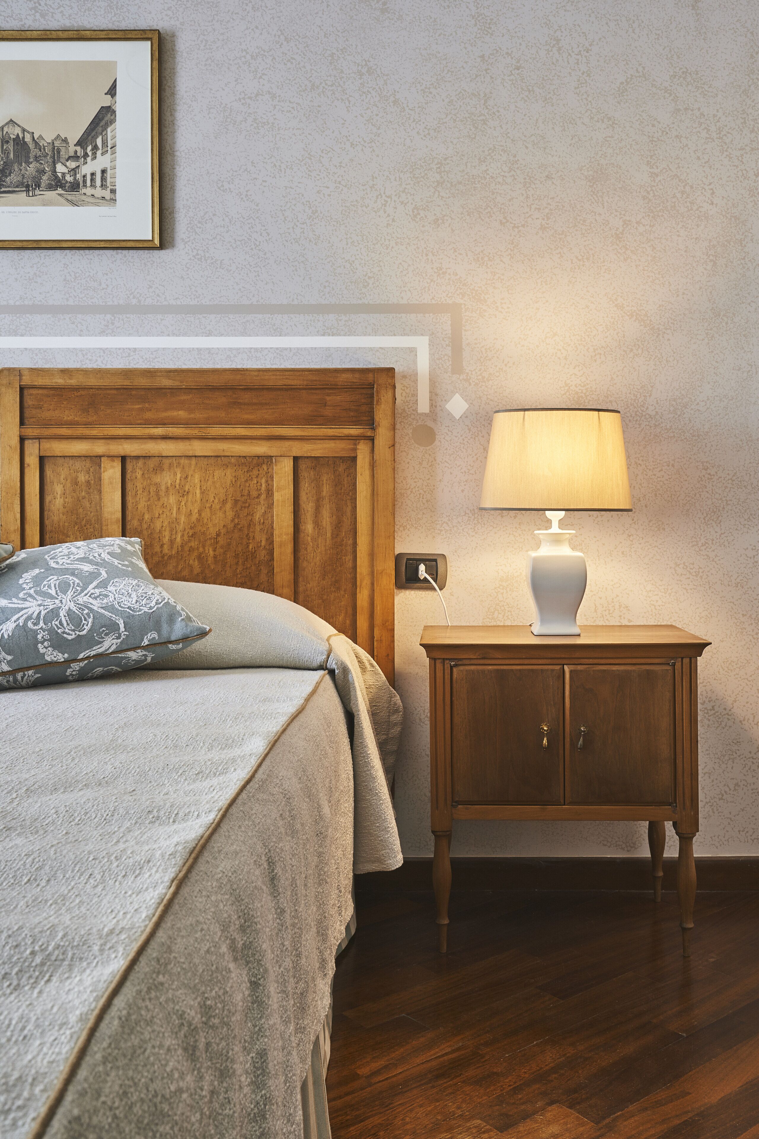 Ein ordentlich bezogenes Bett mit einem hölzernen Kopfteil, ein Nachttisch mit Lampe und ein gerahmtes Bild an der Wand.