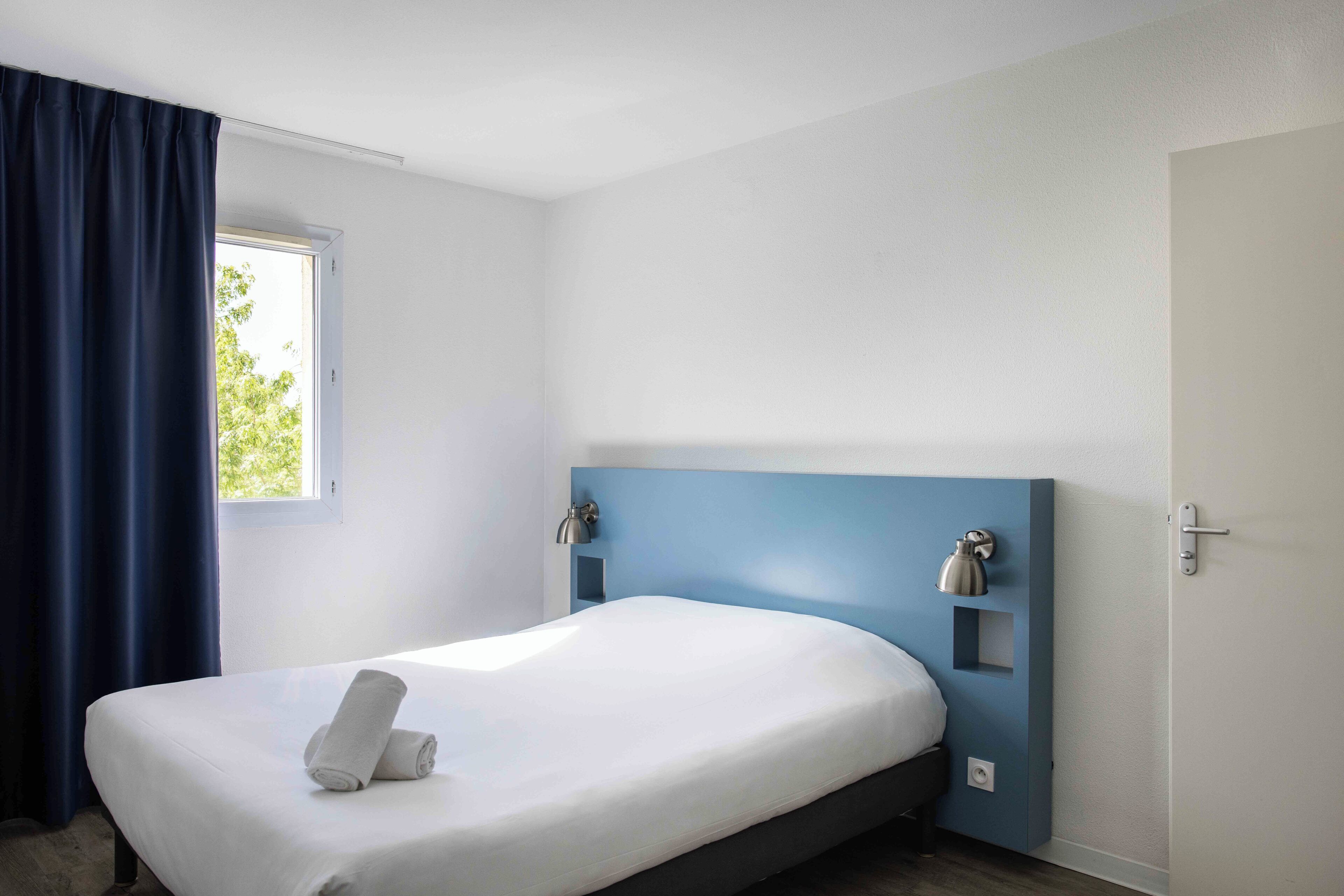 A single bed with a white mattress and a rolled towel, a blue headboard, a wall-mounted lamp, and a window with curtains.