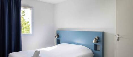 Una cama individual con colchón blanco, una toalla enrollada, cabecera azul, lámpara de pared y una ventana con cortinas.