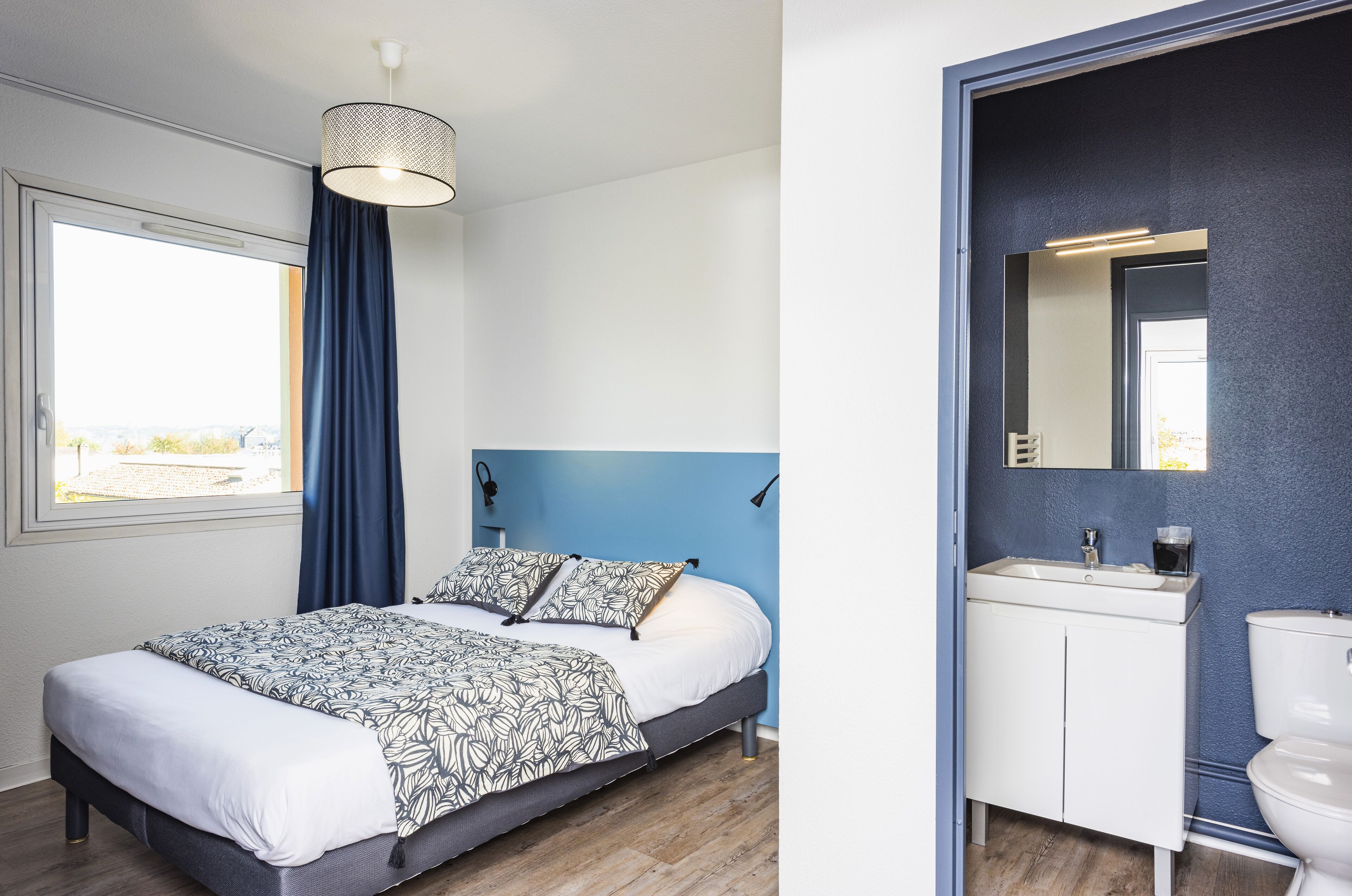 A modern bedroom with a bed, a window with curtains, and a bathroom with a sink and toilet.