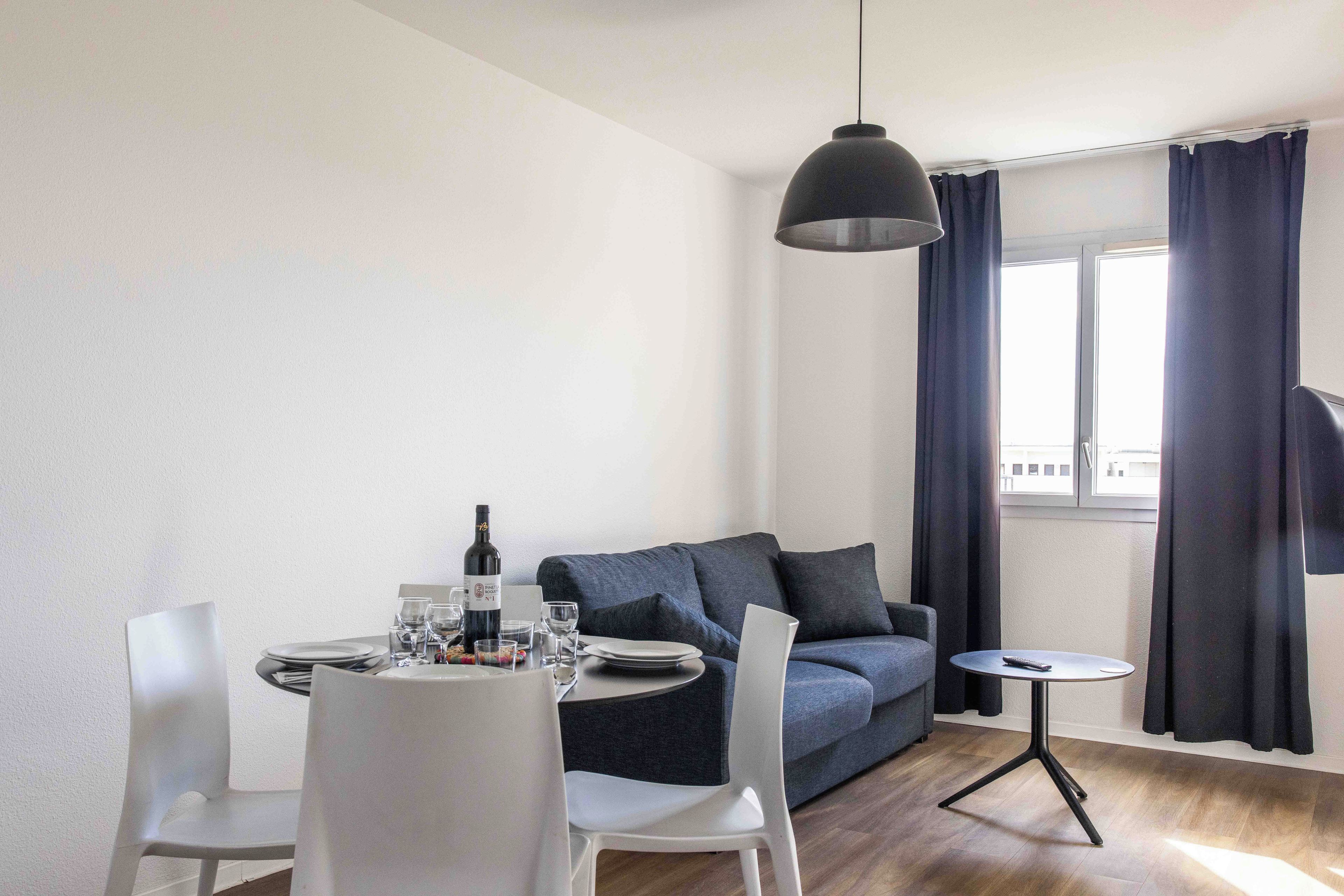 Una sala de estar moderna con una mesa de comedor para dos personas, un sofá, una pequeña mesita auxiliar y un ventanal con cortinas.