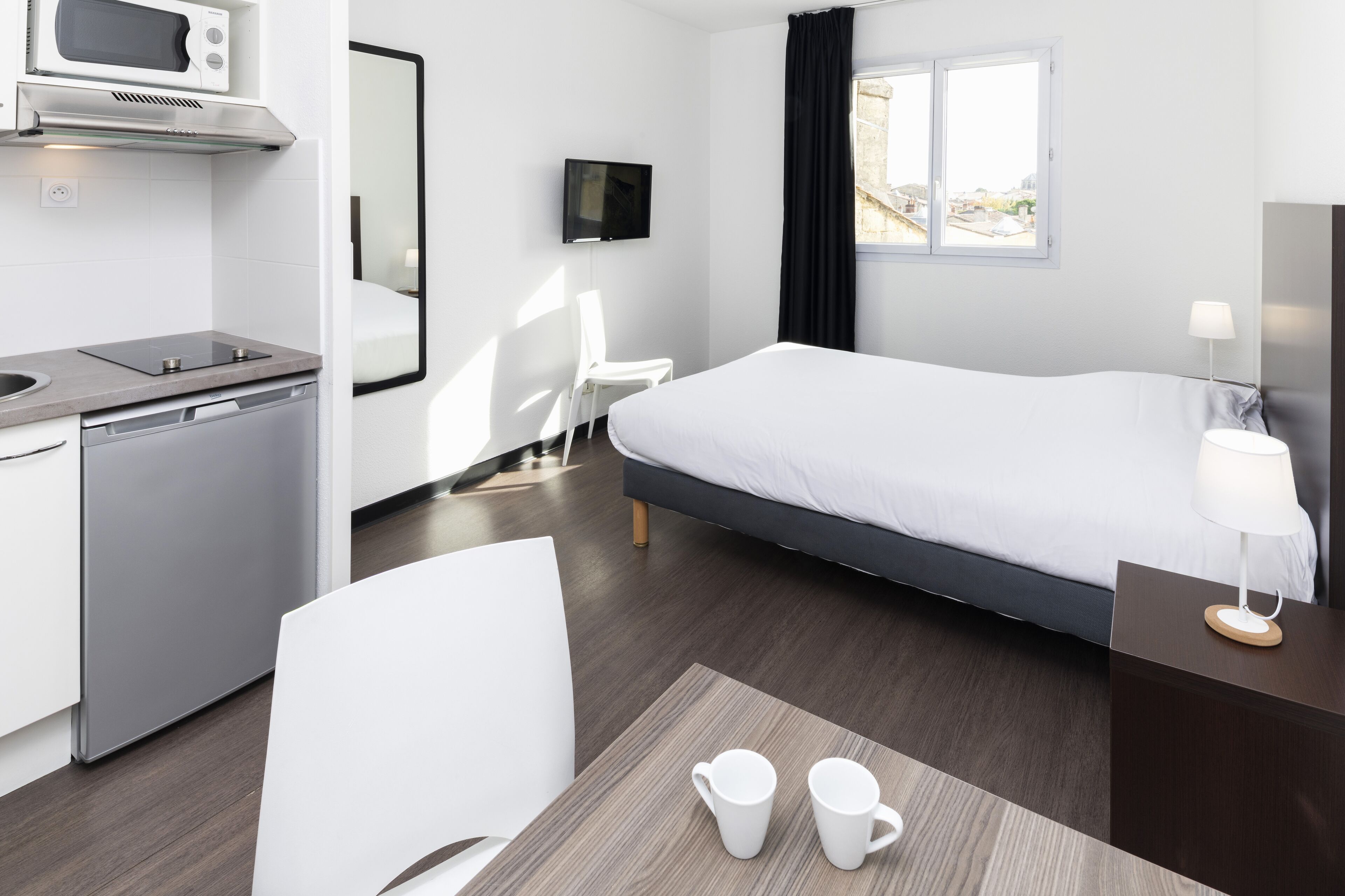 A modern hotel room with a bed, a desk with a chair, a microwave, a TV, and a window with curtains.