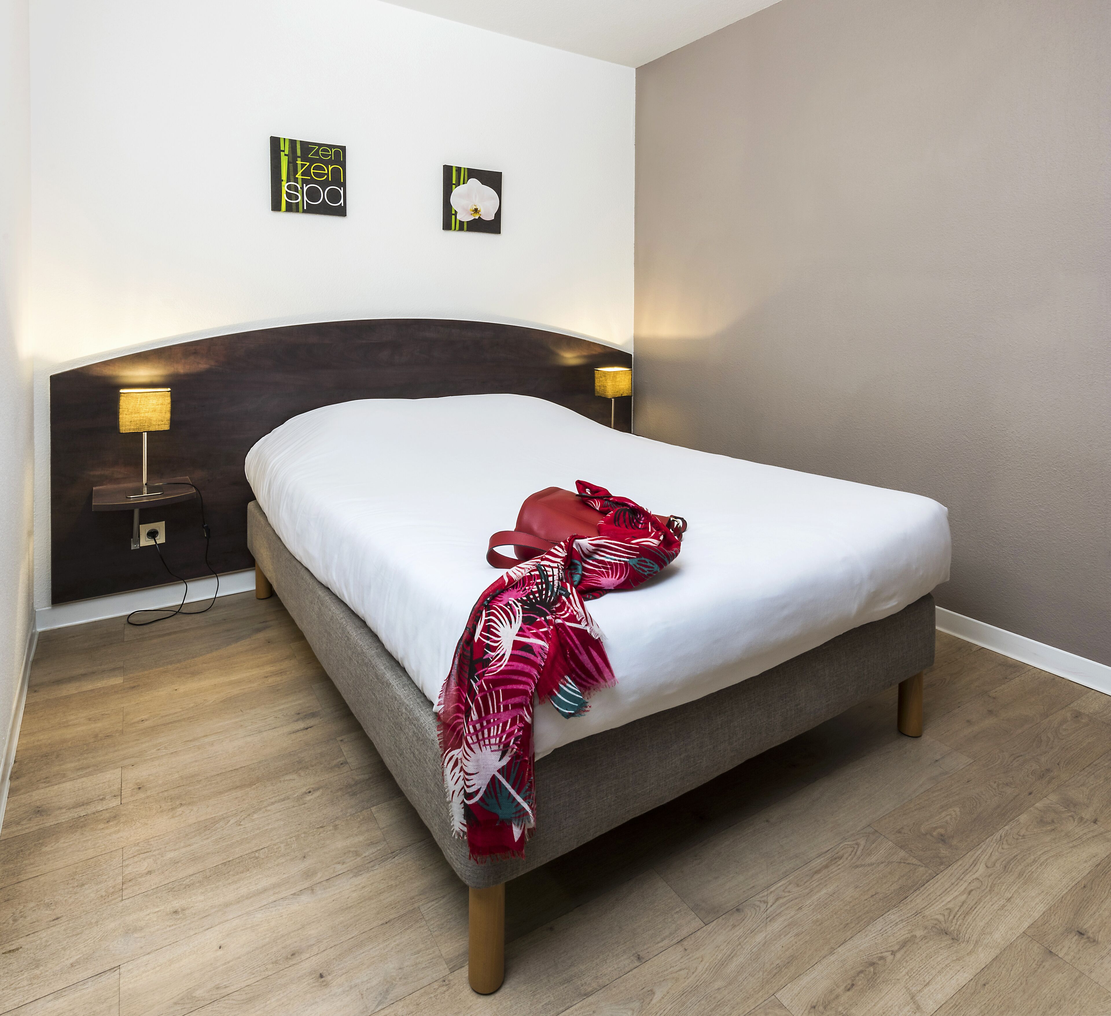 Una habitación de hotel con una cama, cabecera, dos lámparas fijadas a la pared y suelo de madera.