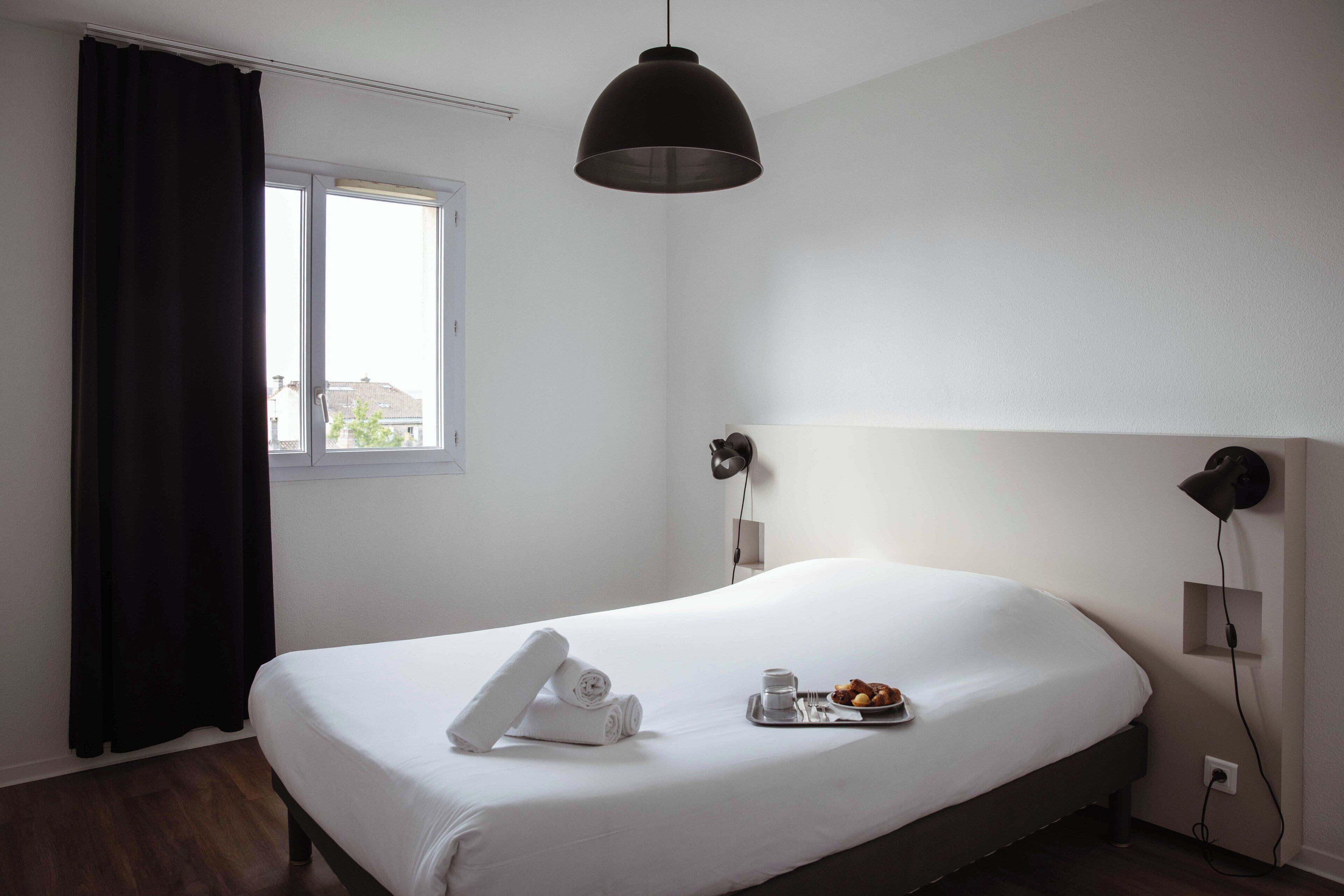 A hotel room with a bed, a window, a wall-mounted lamp, and a tray with breakfast.