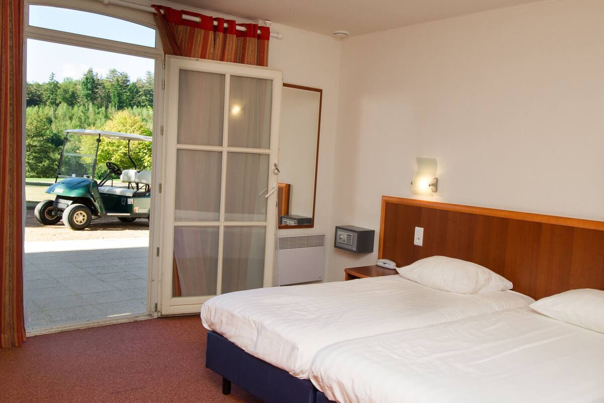 Habitación de hotel con cama, mesita de noche, teléfono y ventana con vista a un carrito de golf y árboles.
