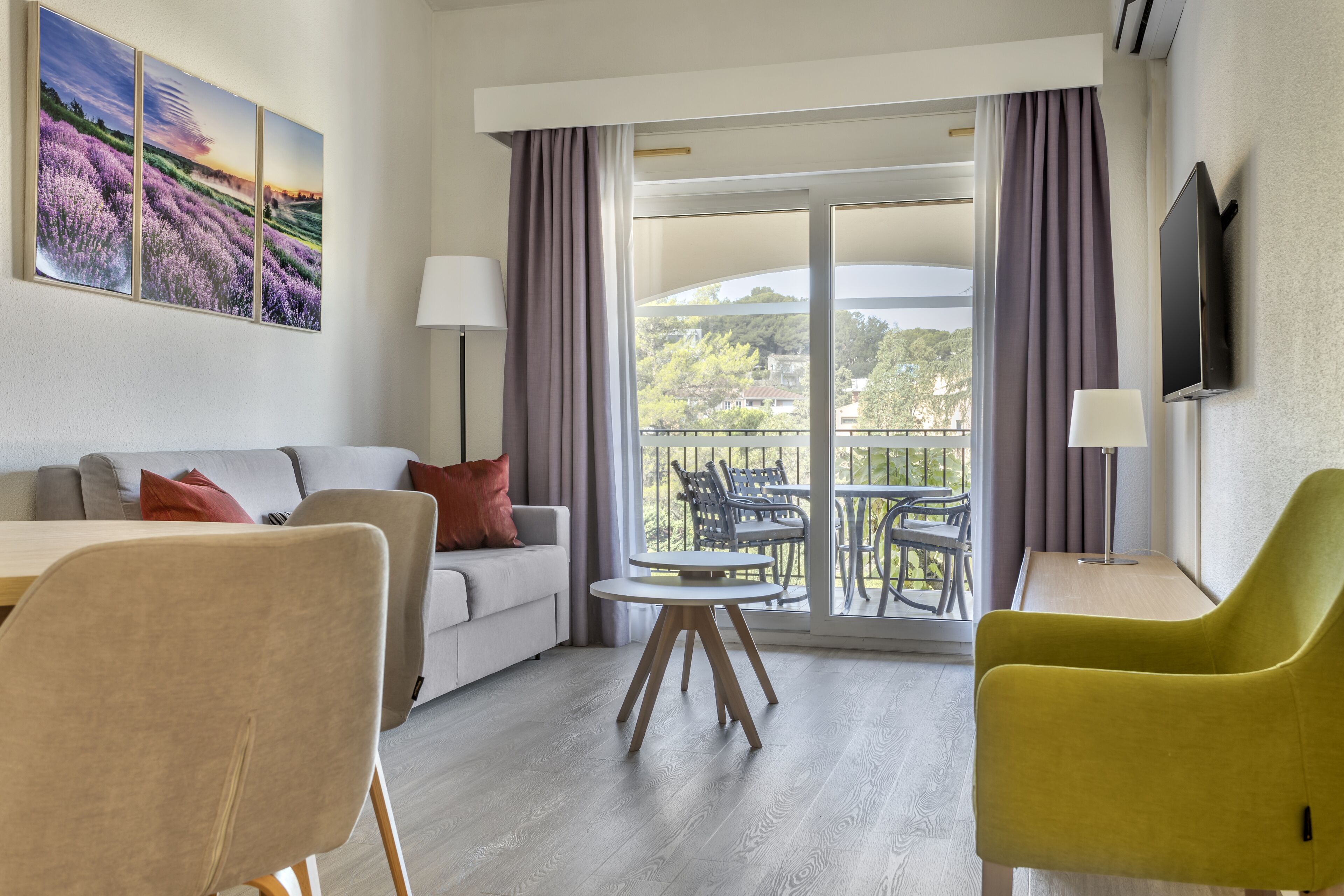A modern hotel room with a balcony, a sofa, a dining table, and a TV.
