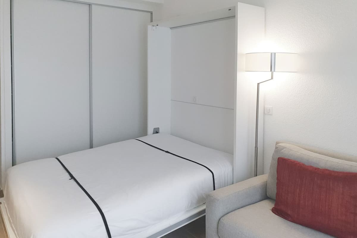 A single bed with a white mattress and black trim, a grey armchair with a red pillow, and a wall-mounted lamp.