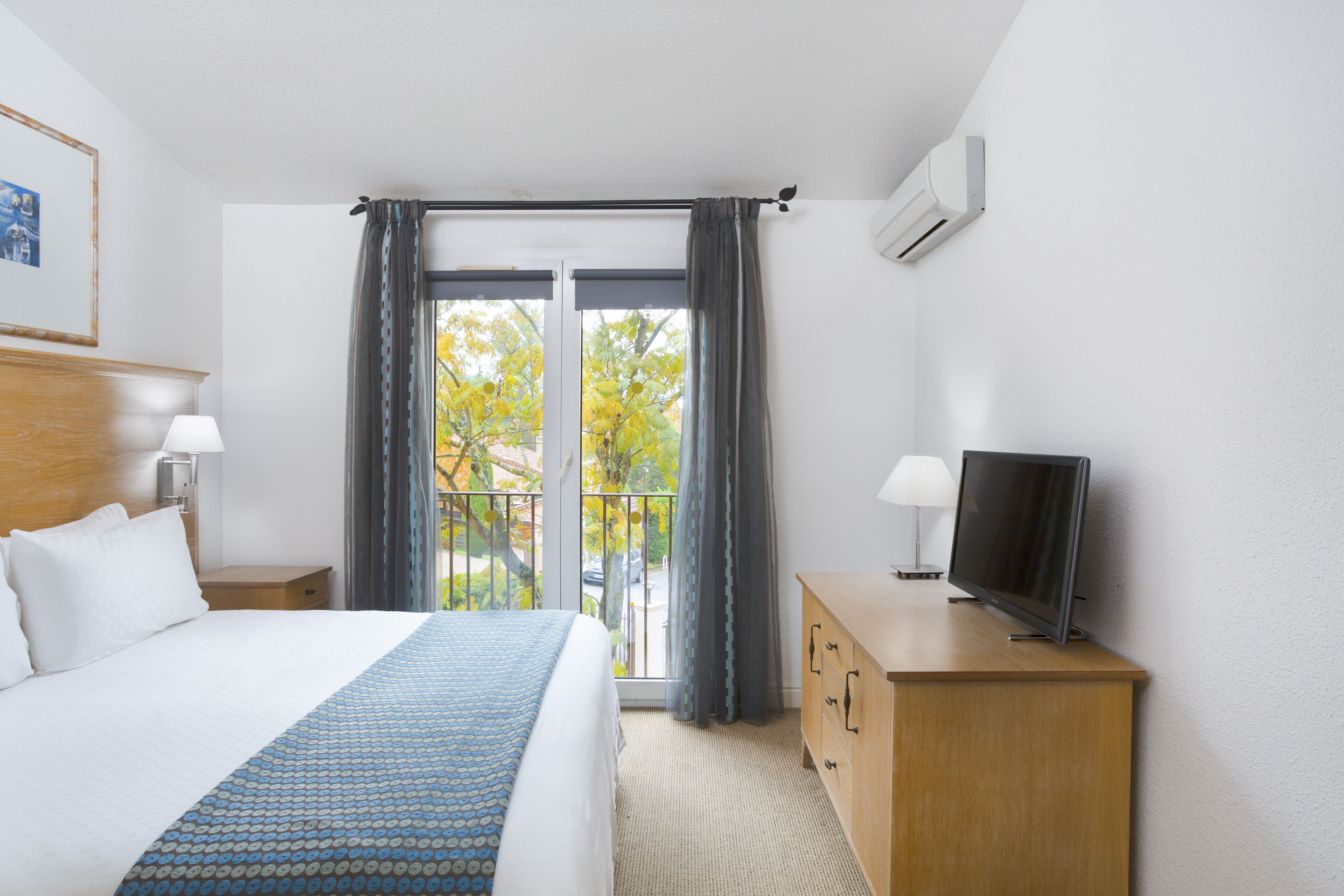 Een hotelkamer met een bed, een televisie op een houten dressoir, een raam met gordijnen en een airconditioner aan de muur.