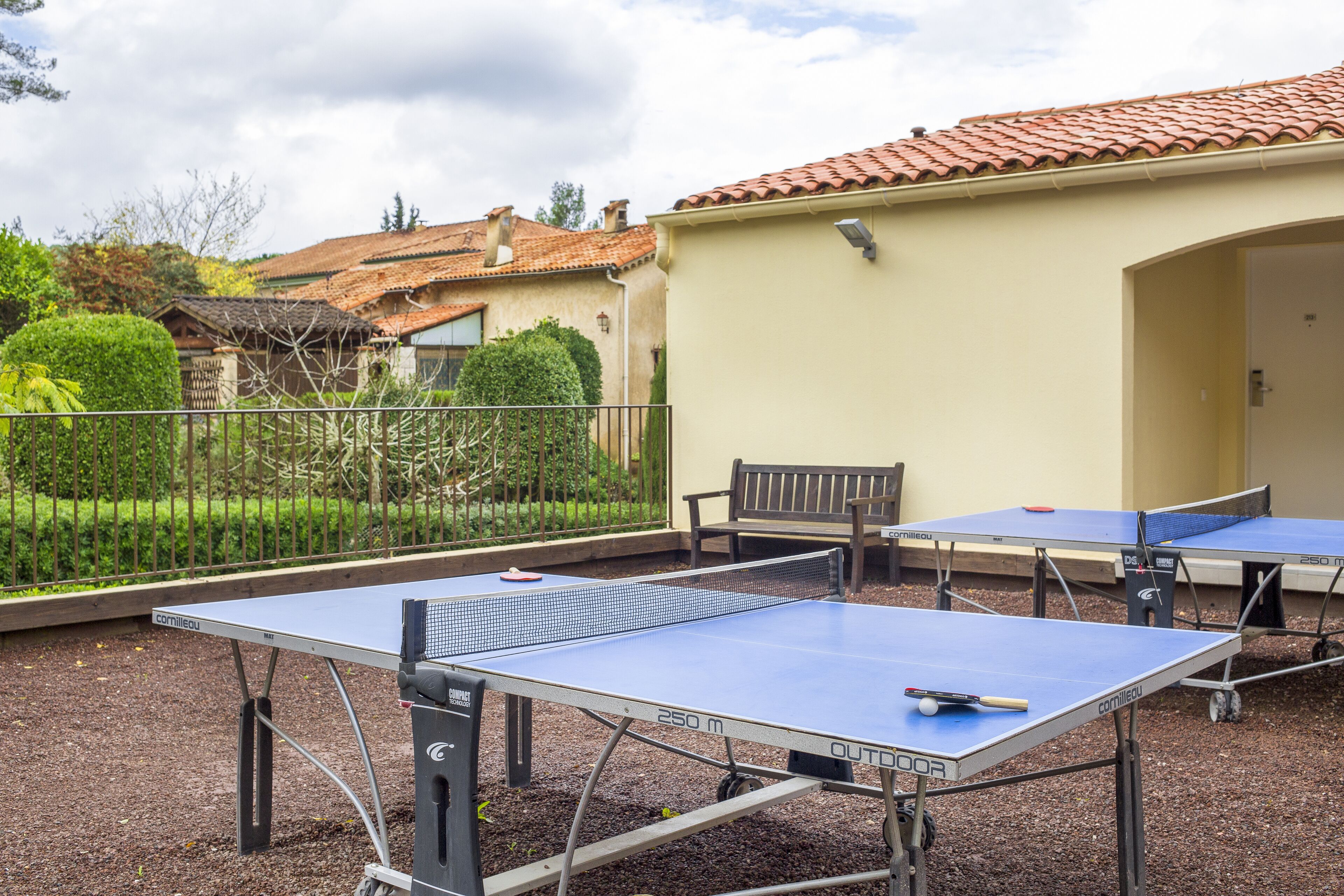 An outdoor table tennis setup with two tables and a bench, surrounded by a fence and greenery.