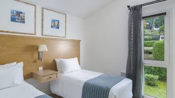 A hotel room with two beds, a wooden headboard, a nightstand, a lamp, and a window with a view of greenery.