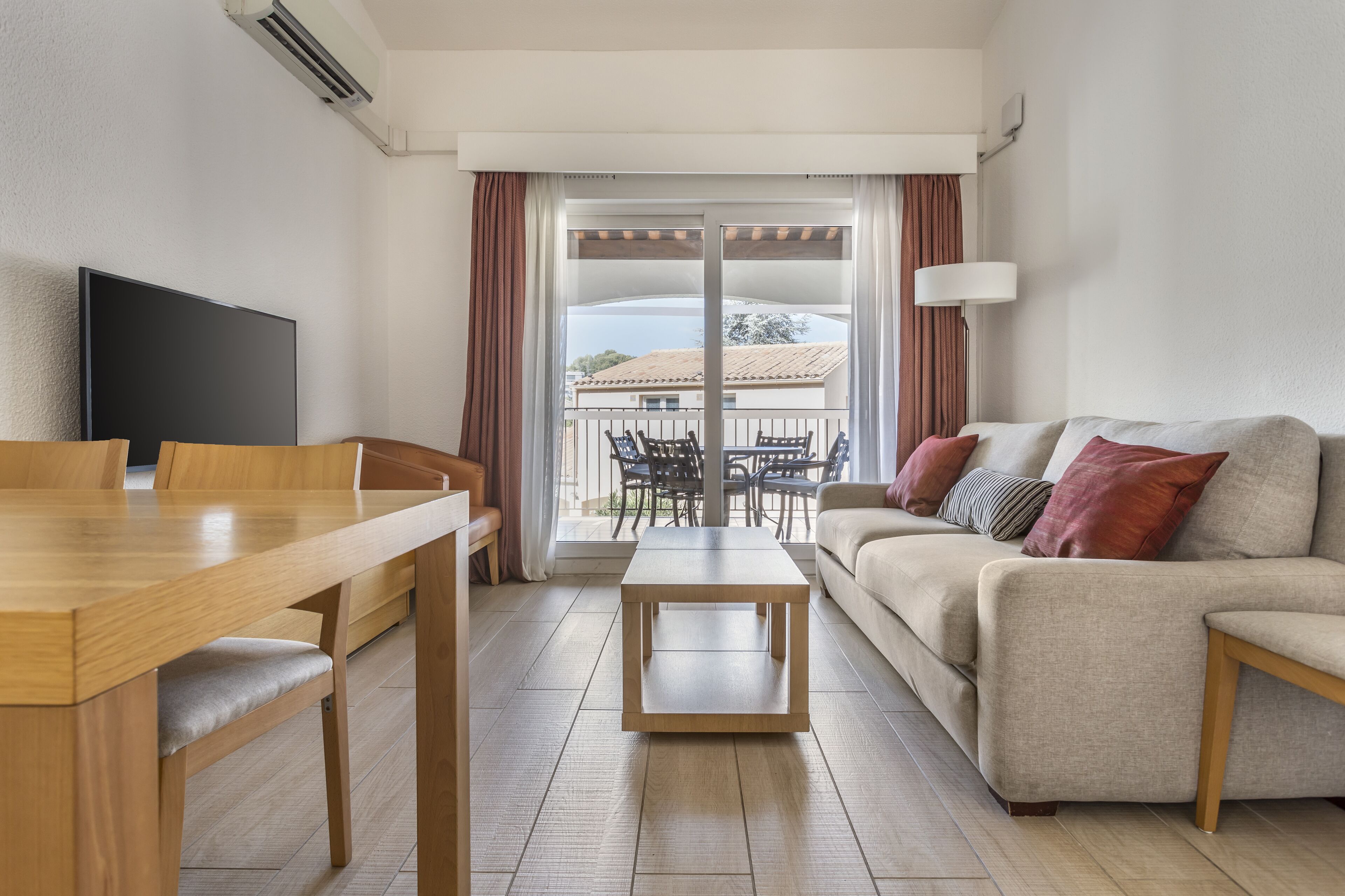 A modern living room with a wooden dining table, a sofa with cushions, a TV, and a balcony with outdoor furniture.
