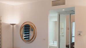 A hotel room with a bed, a round mirror, a floor lamp, and a view into the bathroom. - Hôtel Le Patio de Violette (Uzès)