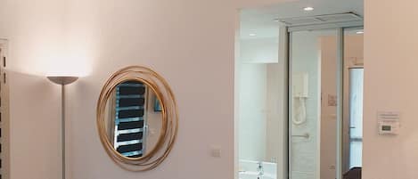A hotel room with a bed, a round mirror, a floor lamp, and a view into the bathroom.