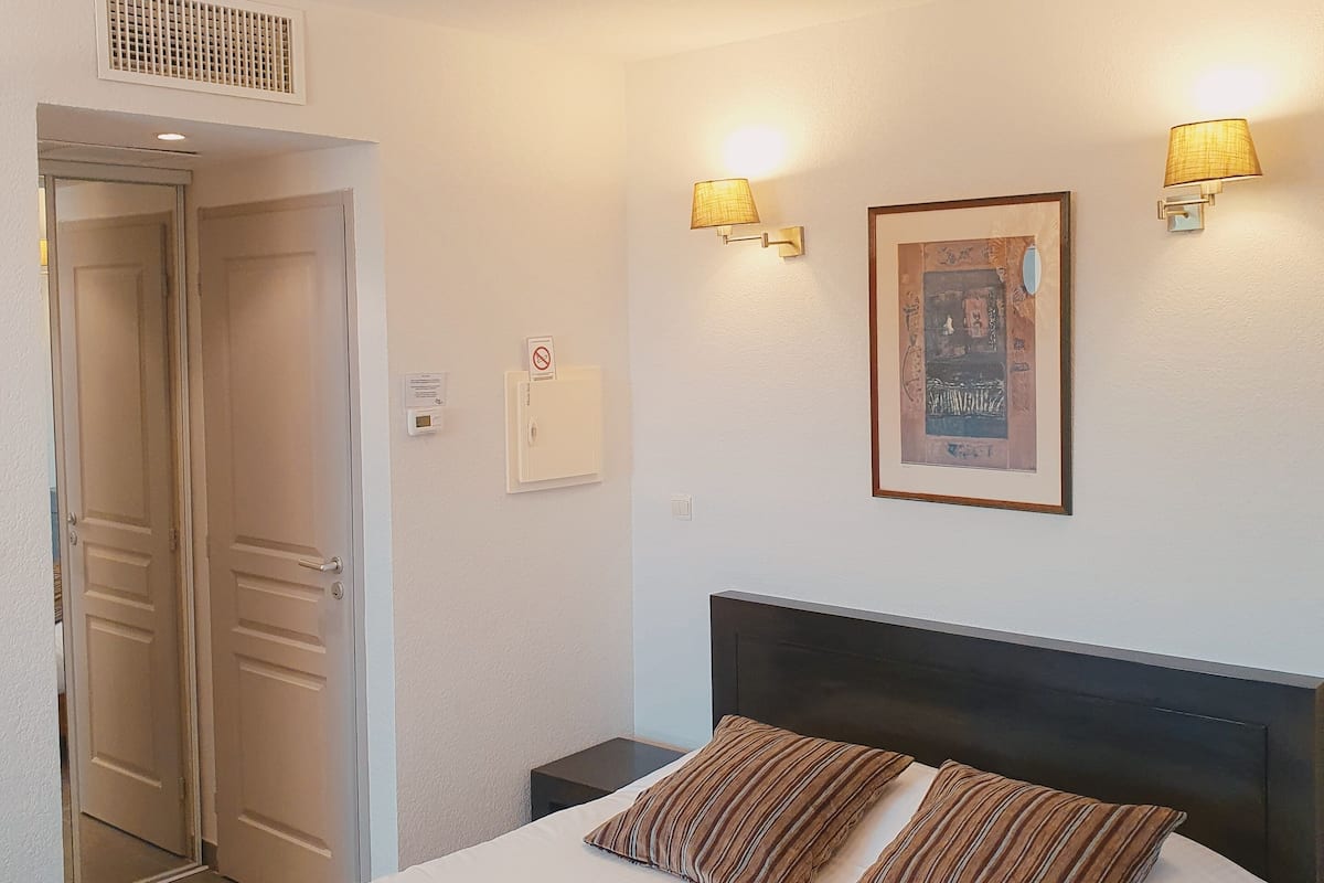 A hotel room with a bed, two bedside lamps, a framed picture on the wall, and a door with a vent above it.