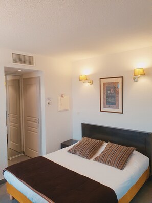 A hotel room with a bed, two bedside lamps, a framed picture on the wall, and a door with a vent above it. - Hôtel Le Patio de Violette (Uzès)