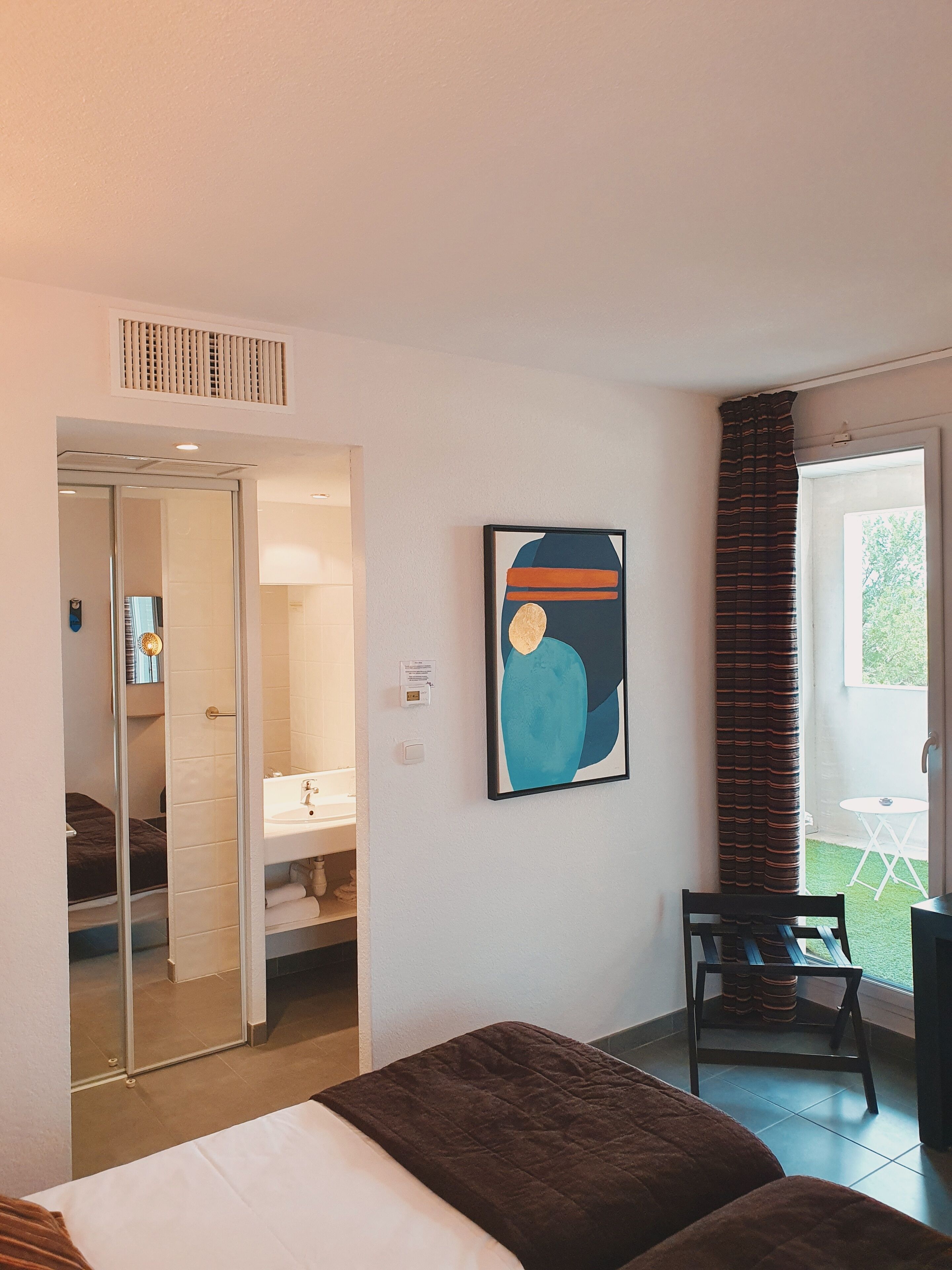 A hotel room with a bed, a bathroom, a painting, and a view of the outdoors.