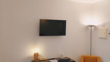 Una habitación de hotel con escritorio, silla, cama, televisor montado en la pared y lámpara de pie.