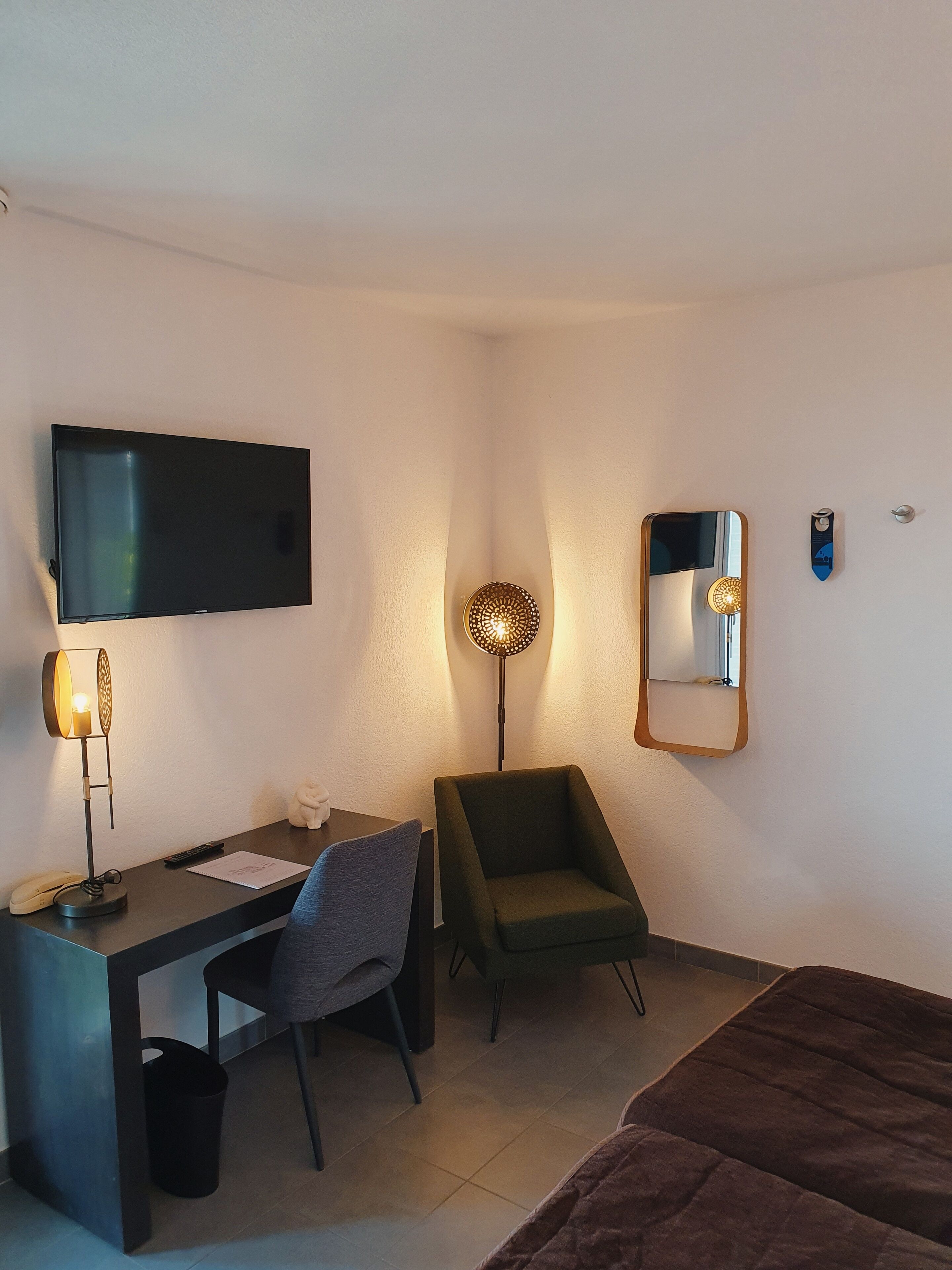 A hotel room with a flat-screen TV, a desk with a chair, a standing lamp, a mirror, and a bed.