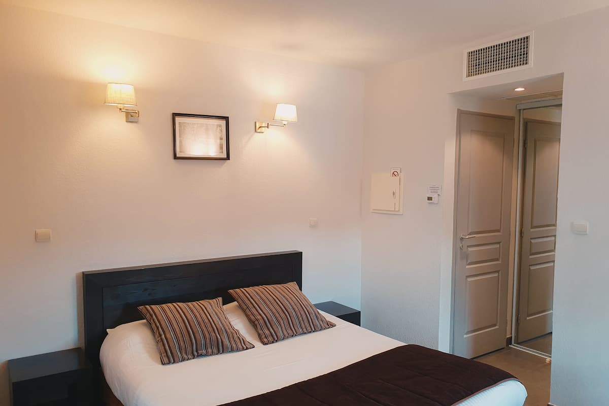 A hotel room with a bed, two wall-mounted lights, a framed picture, a door, and a vent.