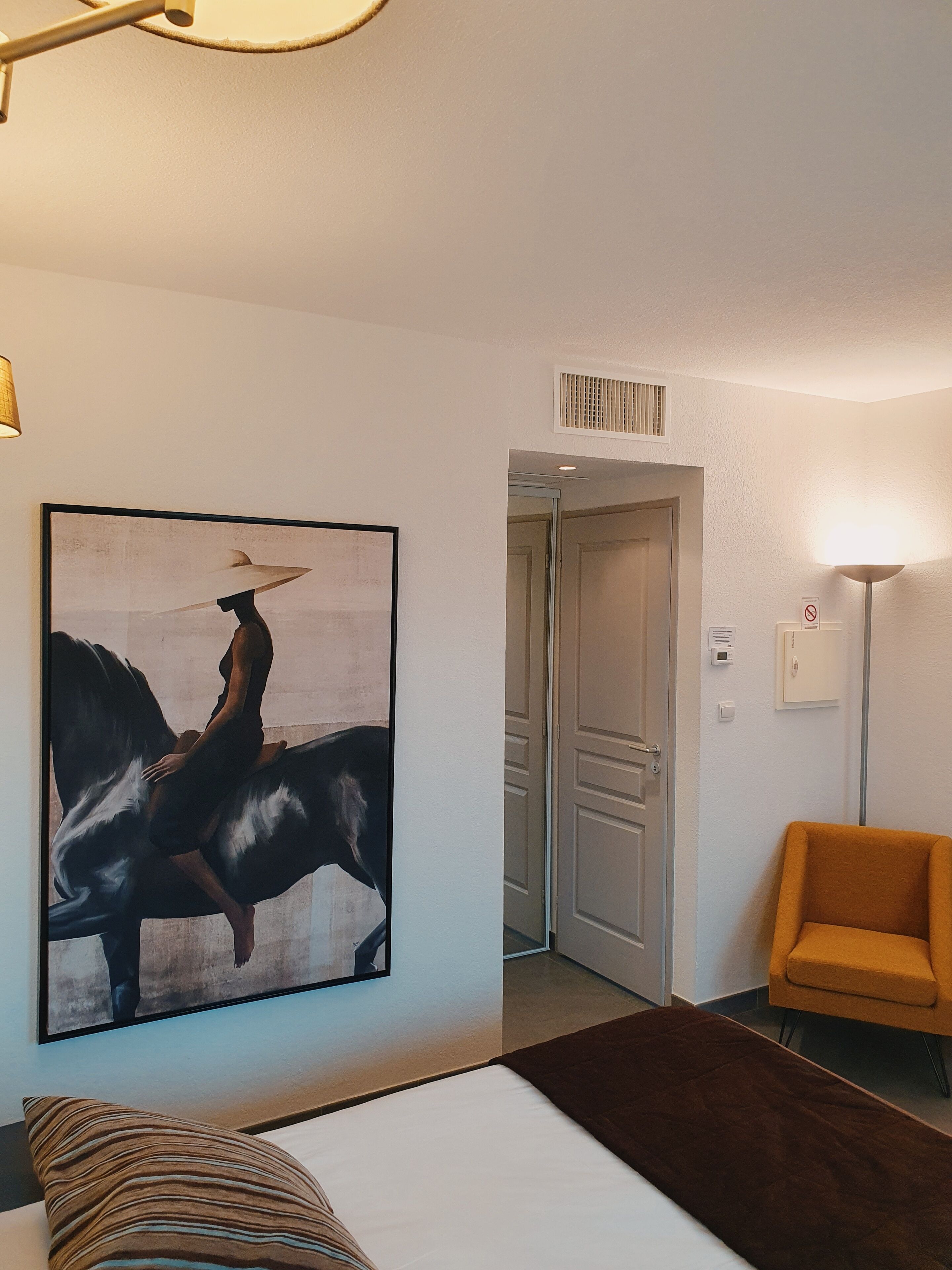 A hotel room with a bed, a large painting of horses, a door, and a chair.