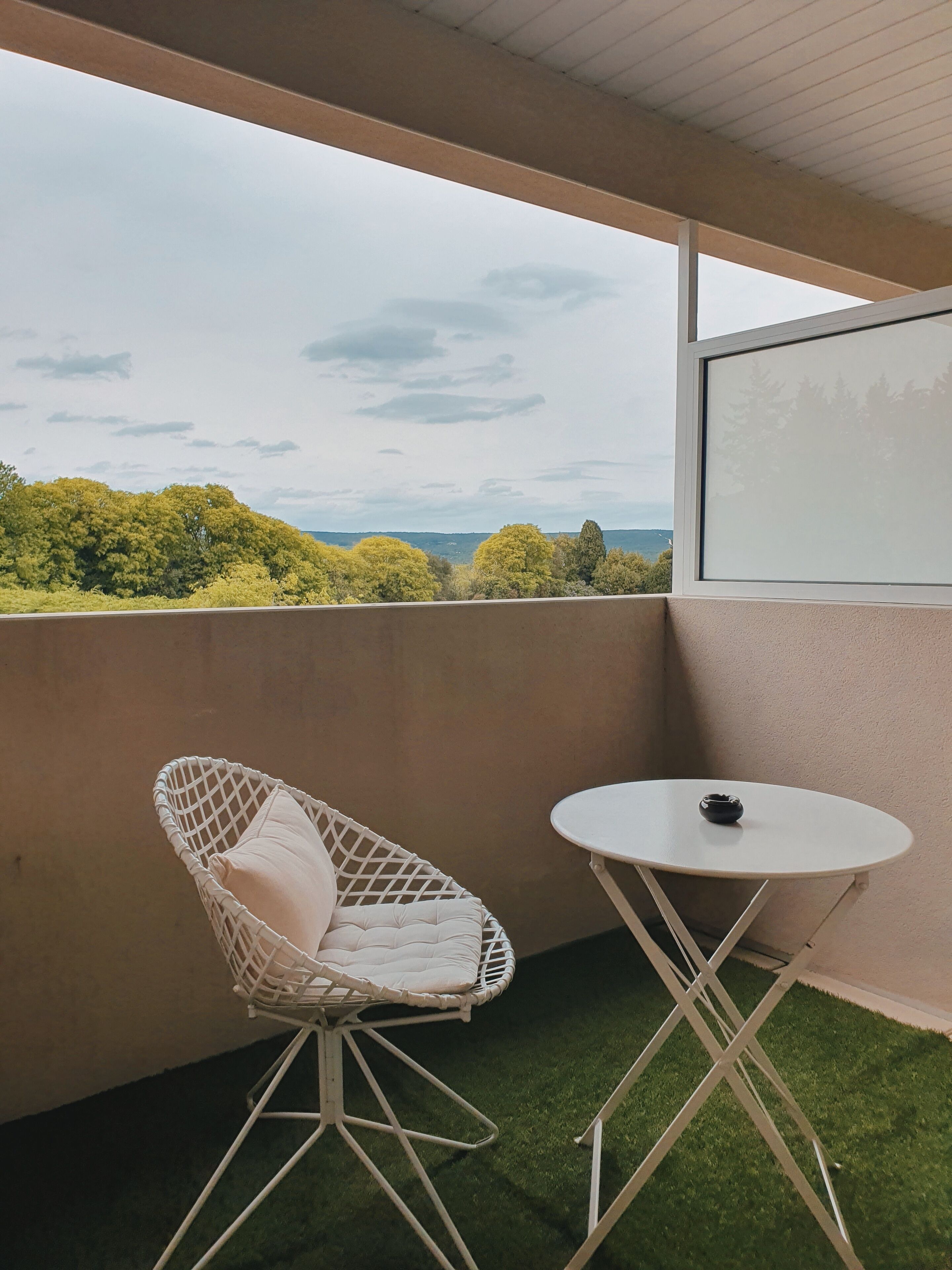 Un balcón con una silla de mimbre blanca y una pequeña mesa redonda, que ofrece vistas a árboles y un paisaje distante.