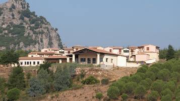 A cluster of Mediterranean-style buildings with terracotta roofs, set against a backdrop of a rocky mountain and dense pine forest.