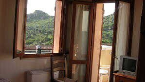 Un dormitorio con una cama, una silla, un aparador y un televisor. Hay ventanas con cortinas y vistas a un paisaje verde.