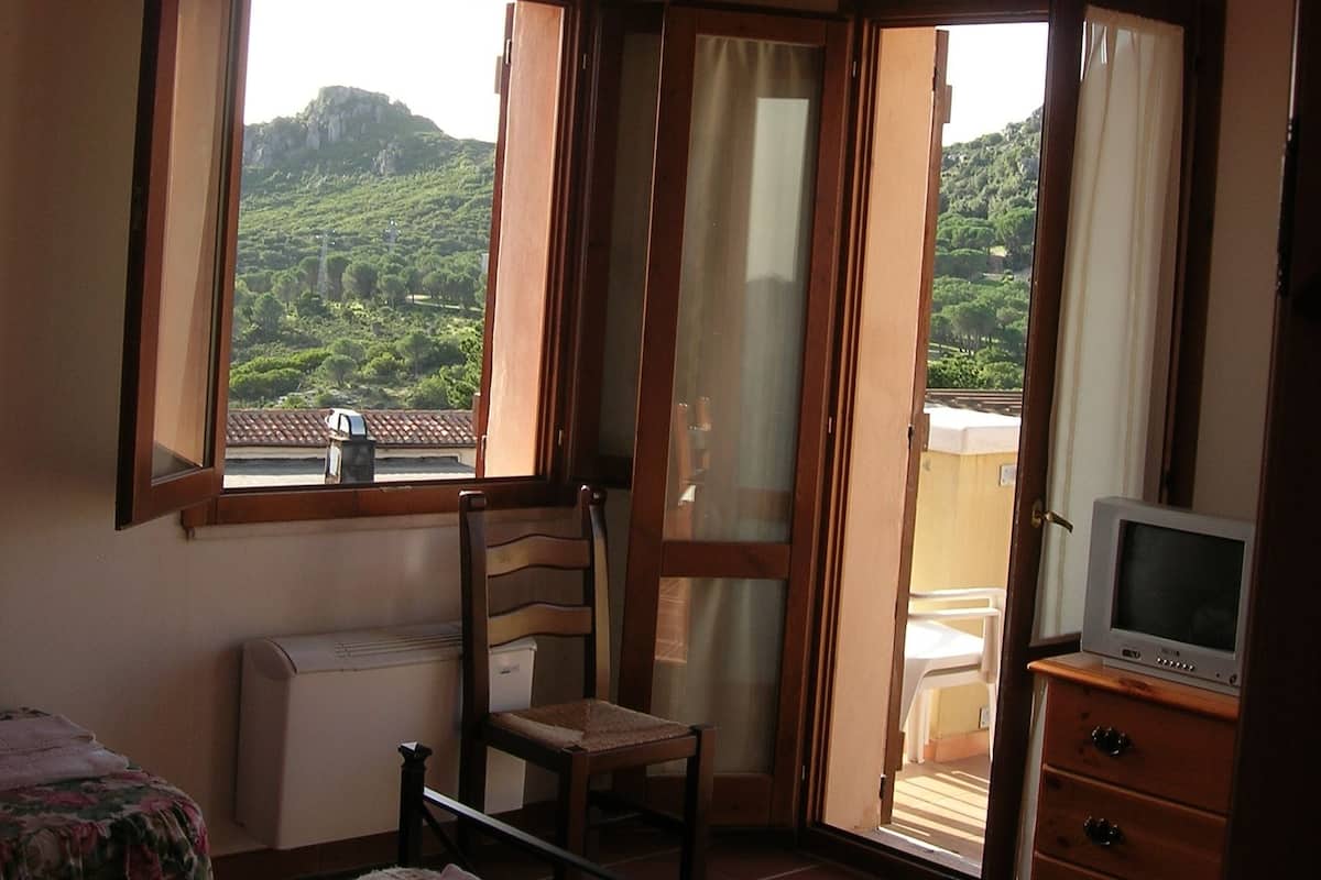 Ein Schlafzimmer mit Bett, Stuhl, Kommode und Fernseher. Es gibt Fenster mit Vorhängen und einen Blick auf eine grüne Landschaft.
