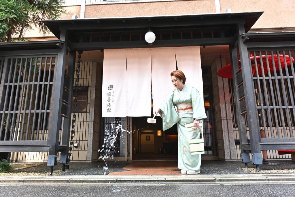 Una persona vestida con traje tradicional frente a la entrada de un edificio con un gran cartel y una puerta japonesa tradicional.