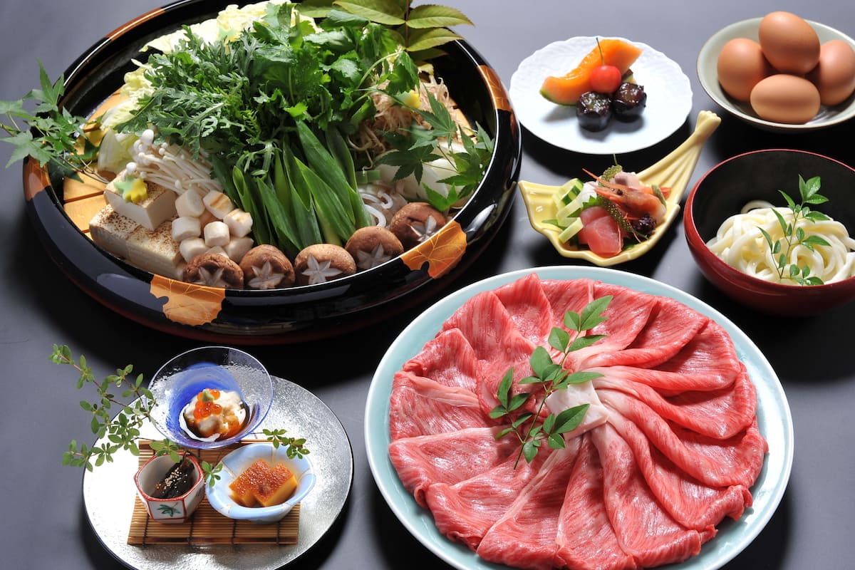 A variety of food items including a bowl of noodles, a plate of raw meat, and several small dishes with condiments.