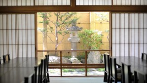 Een traditionele Japanse kamer met tatami vloer, een lage tafel met stoelen en uitzicht op een tuin met een stenen lantaarn en bamboehek.'