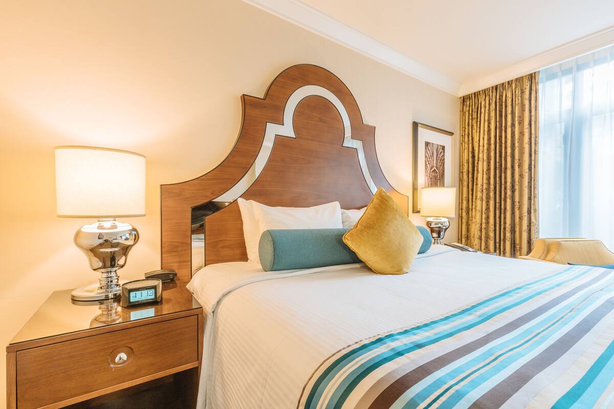 A hotel room with a large bed, a wooden headboard, a nightstand with a lamp, and a window with curtains.