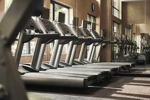 Varias caminadoras alineadas en un gimnasio con amplias ventanas y un reloj en la pared.
