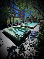 A pool area with two rectangular pools, surrounded by lounge chairs and a brick building in the background.