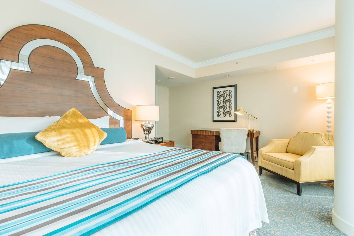 A hotel room with a large bed, a wooden headboard, a striped bedspread, a yellow pillow, a chair, a desk with a lamp, and a framed artwork on the wall.