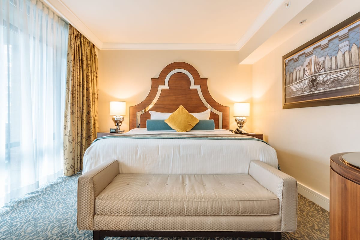 A hotel room with a large bed, two bedside lamps, a headboard with a decorative arch, a sofa, and a painting on the wall.