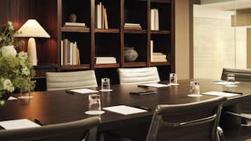 A conference room with a dark wooden table, white chairs, and a bookshelf in the background.