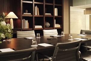 A conference room with a dark wooden table, white chairs, and a bookshelf in the background.