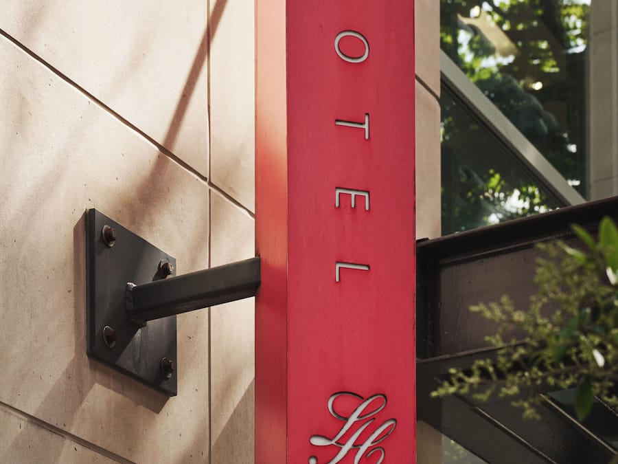 Un letrero rojo de hotel con el nombre 'HOTEL' y un logotipo, sujeto a una pared con un soporte metálico.