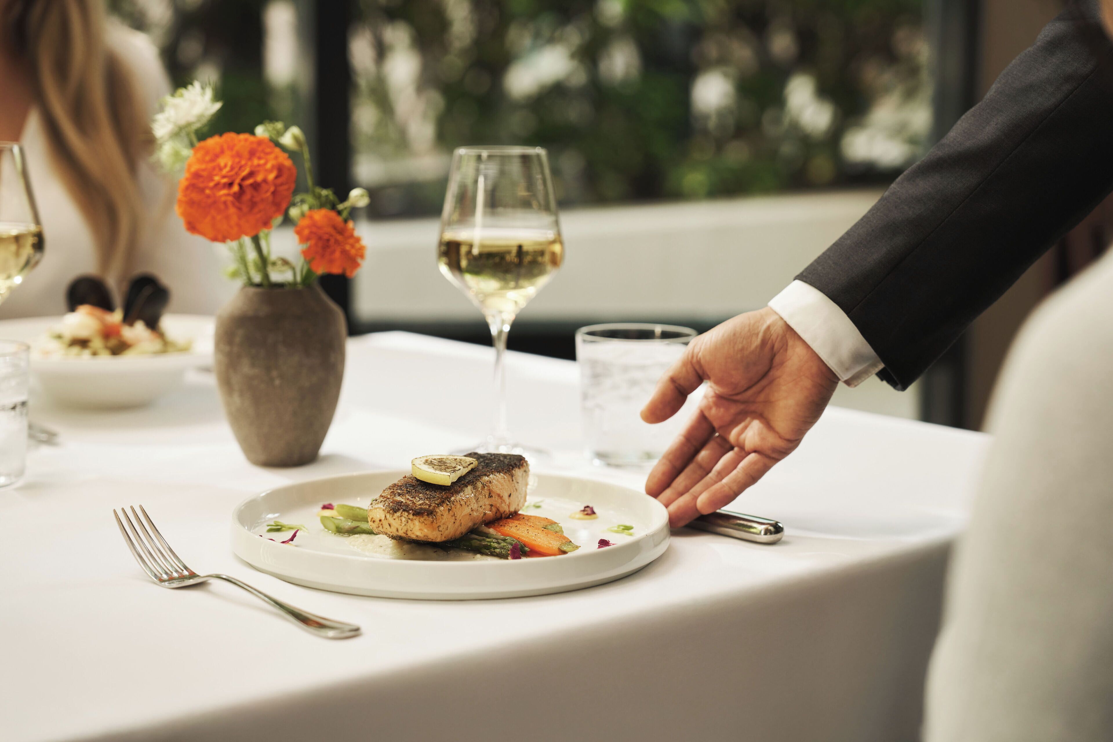 Una mesa de comedor con un plato de comida, una copa de vino blanco y un jarrón con flores.