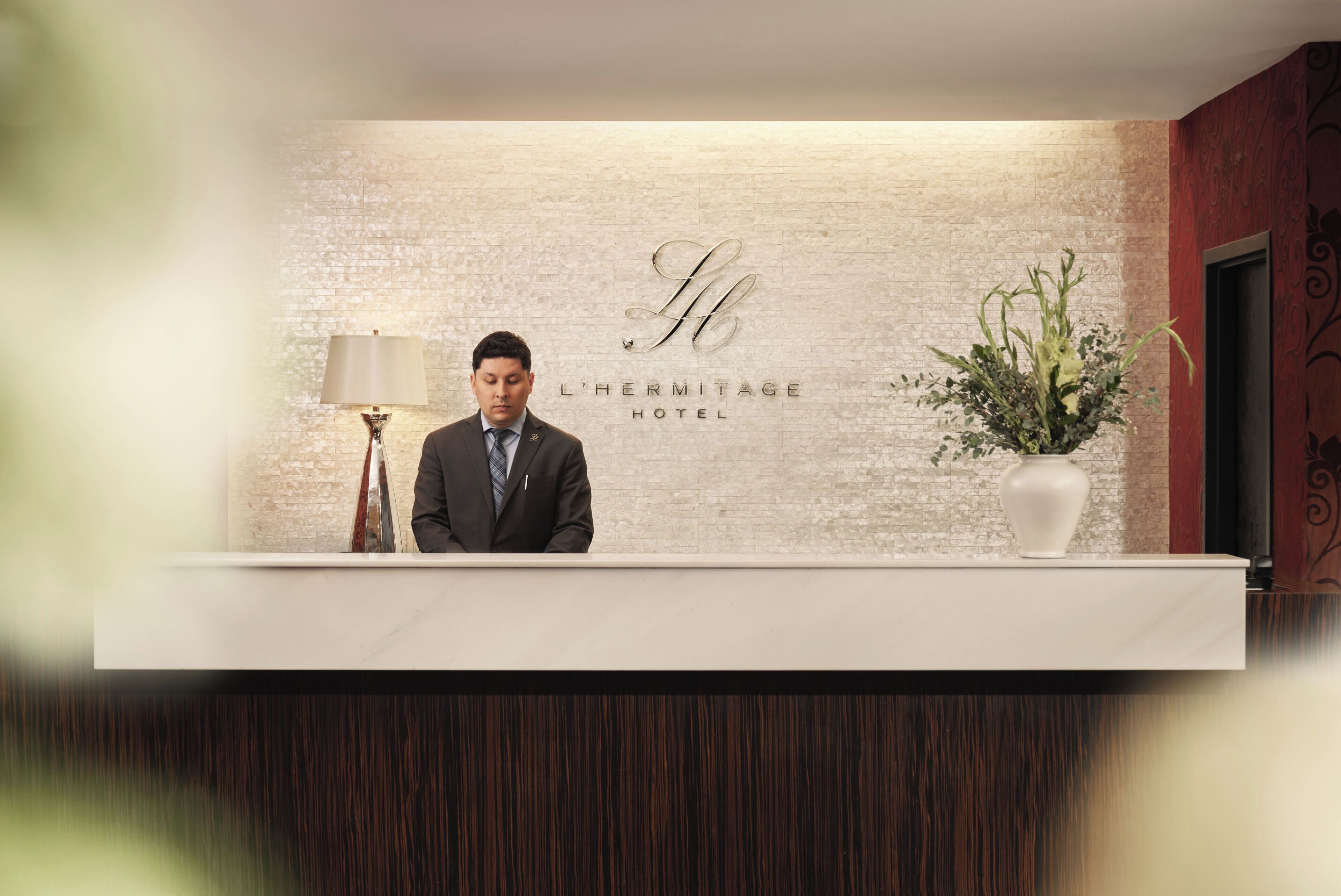 A hotel reception desk with a brick wall background, a sign reading 'L'HERMITAGE HOTEL', and a potted plant to the right.