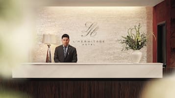 A hotel reception desk with a brick wall background, a sign reading 'L'HERMITAGE HOTEL', and a potted plant to the right.