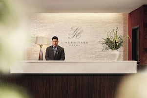 A hotel reception desk with a brick wall background, a sign reading 'L'HERMITAGE HOTEL', and a potted plant to the right.