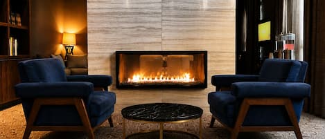 A modern lounge area with a fireplace, two blue armchairs, a round coffee table, and a patterned carpet.