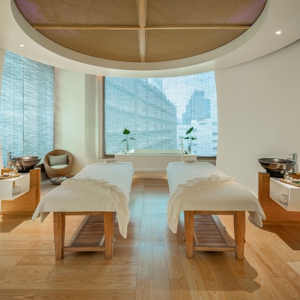A spa room with two treatment beds, wooden flooring, a large window with blinds, and a ceiling with a woven design.