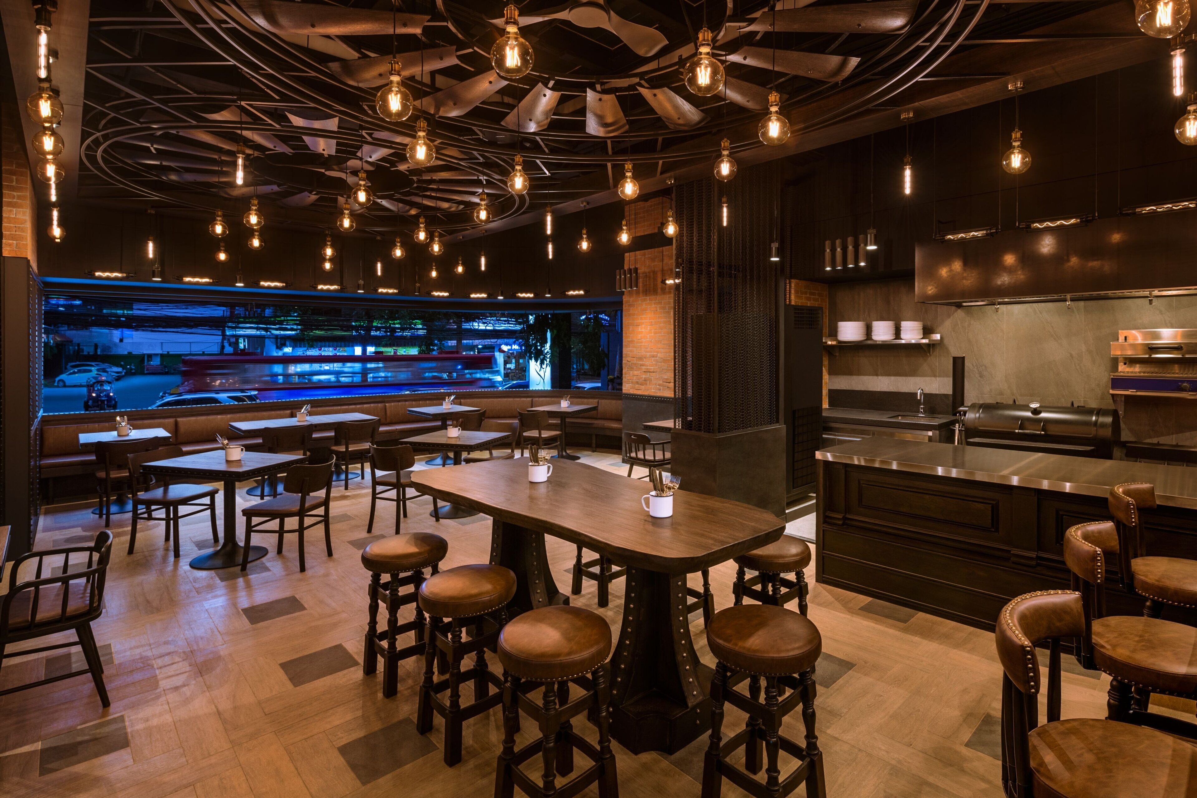 A restaurant with wooden tables and stools, a bar area, and a kitchen visible through the glass wall.