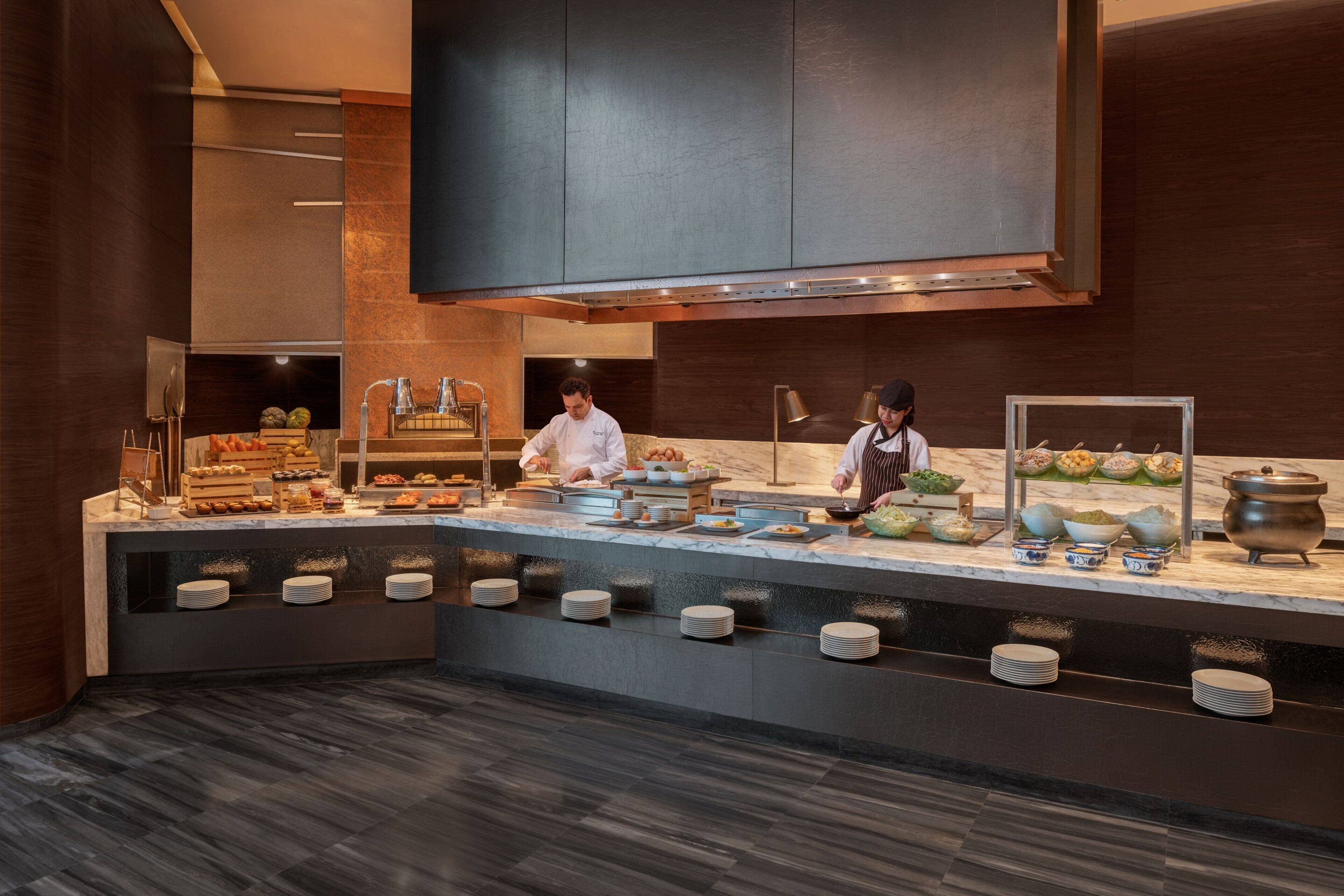 A modern kitchen with a marble countertop, chef preparing food, and a buffet area with various dishes and bowls.